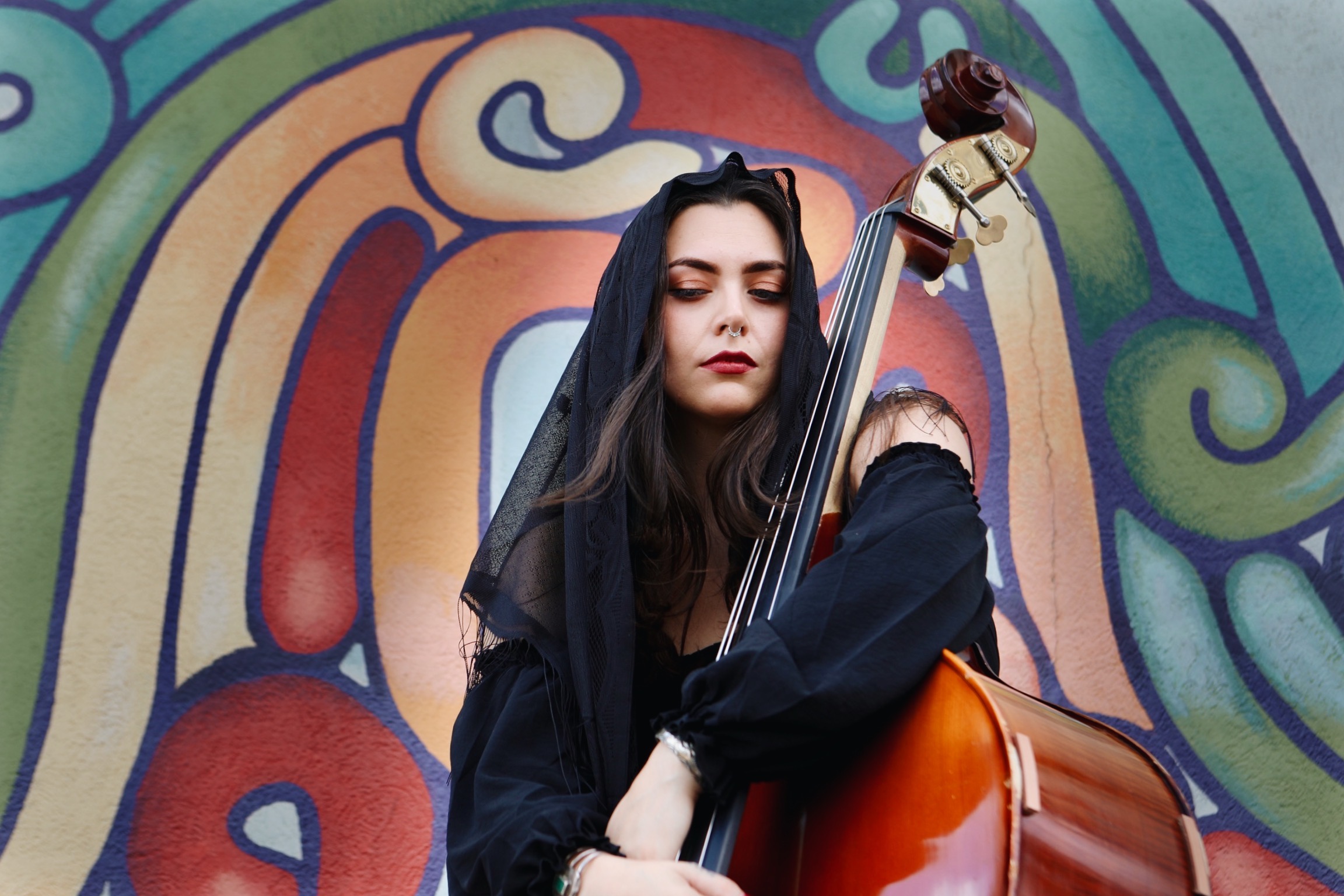 Young woman with long dark hair and dark clothing holding a cello in front of a colorful mural background.