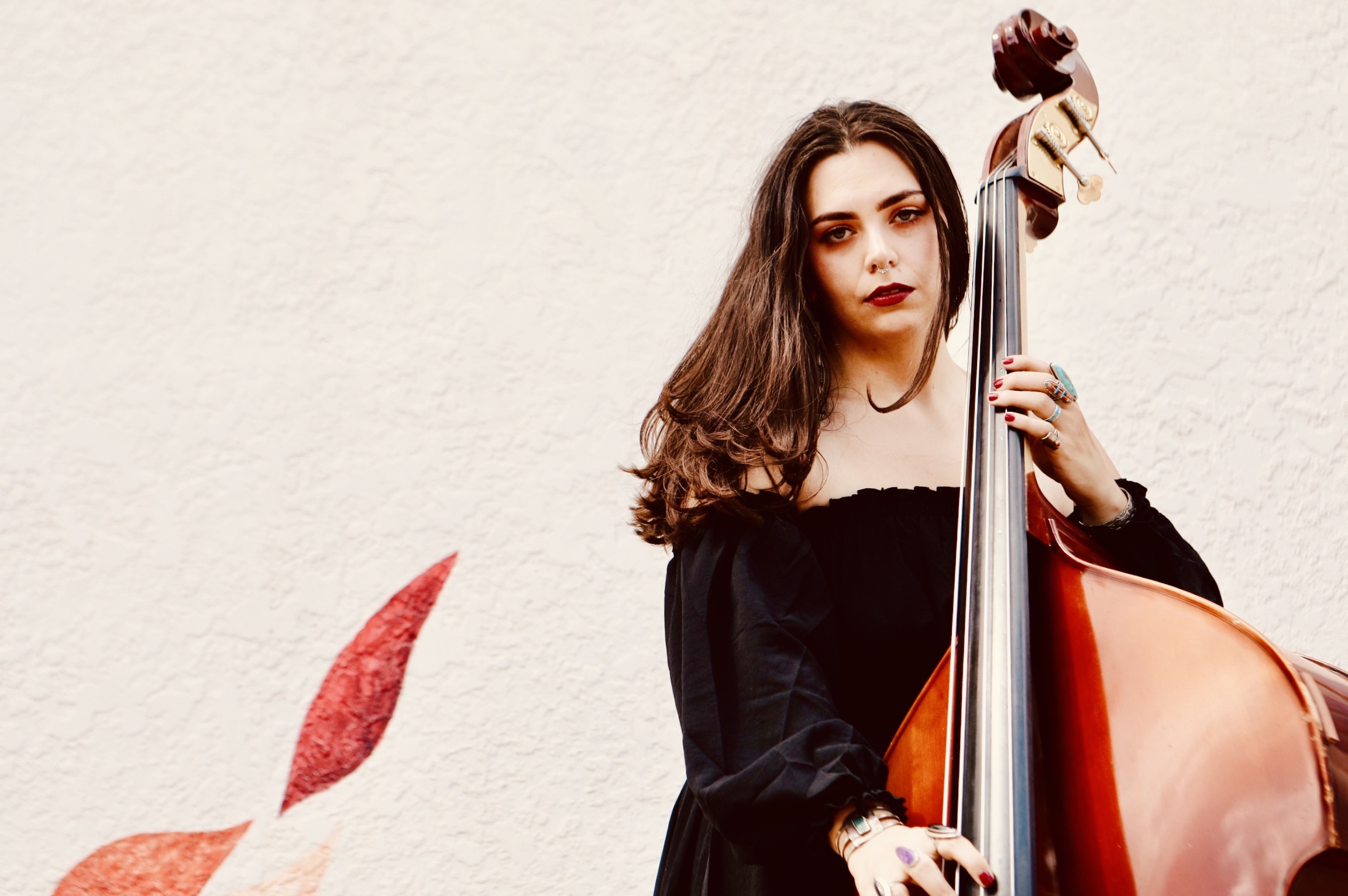 Young woman with long brown hair in a black dress playing a double bass against a light textured wall.