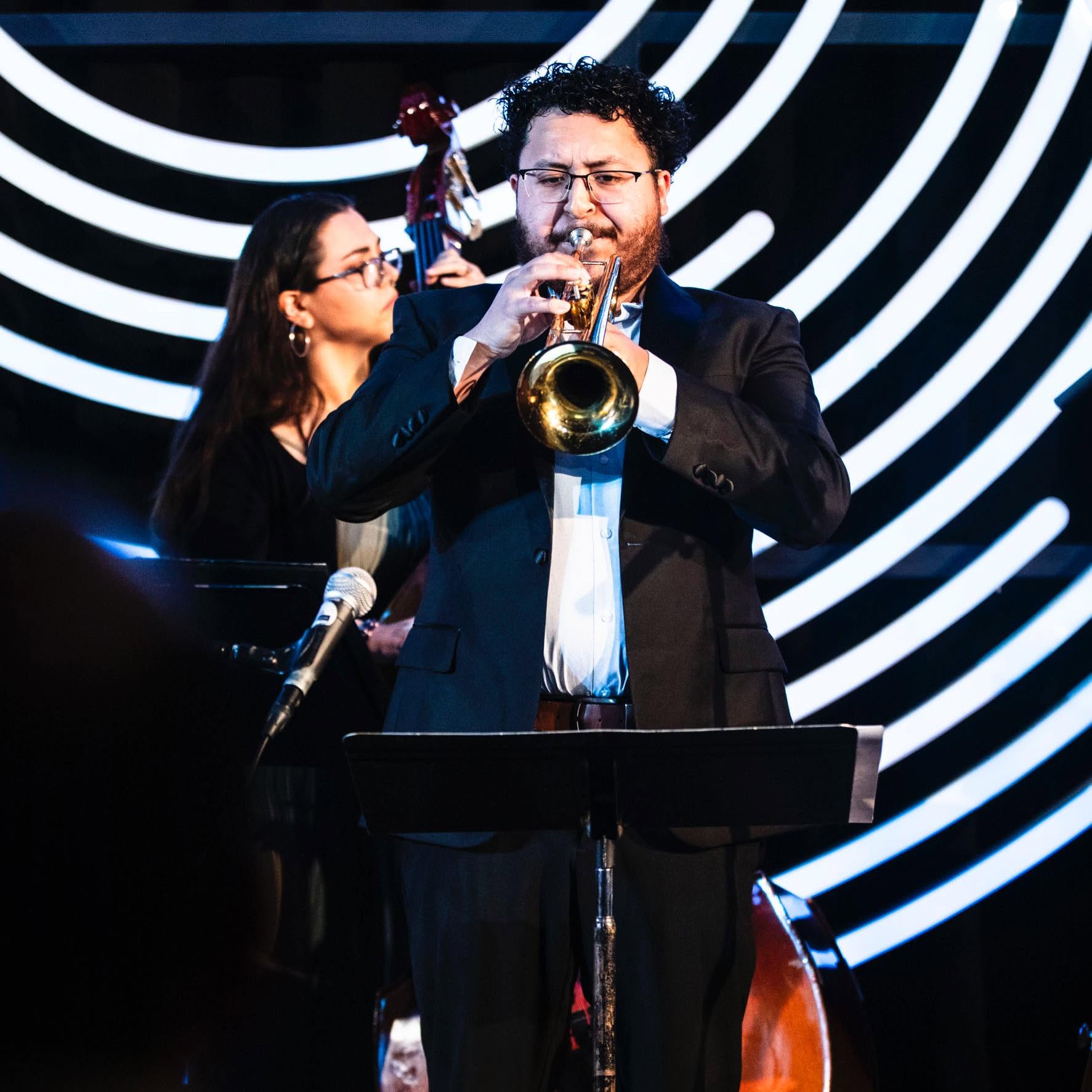 A man playing a trumpet on stage with a woman in the background, against a black and white spiral backdrop.