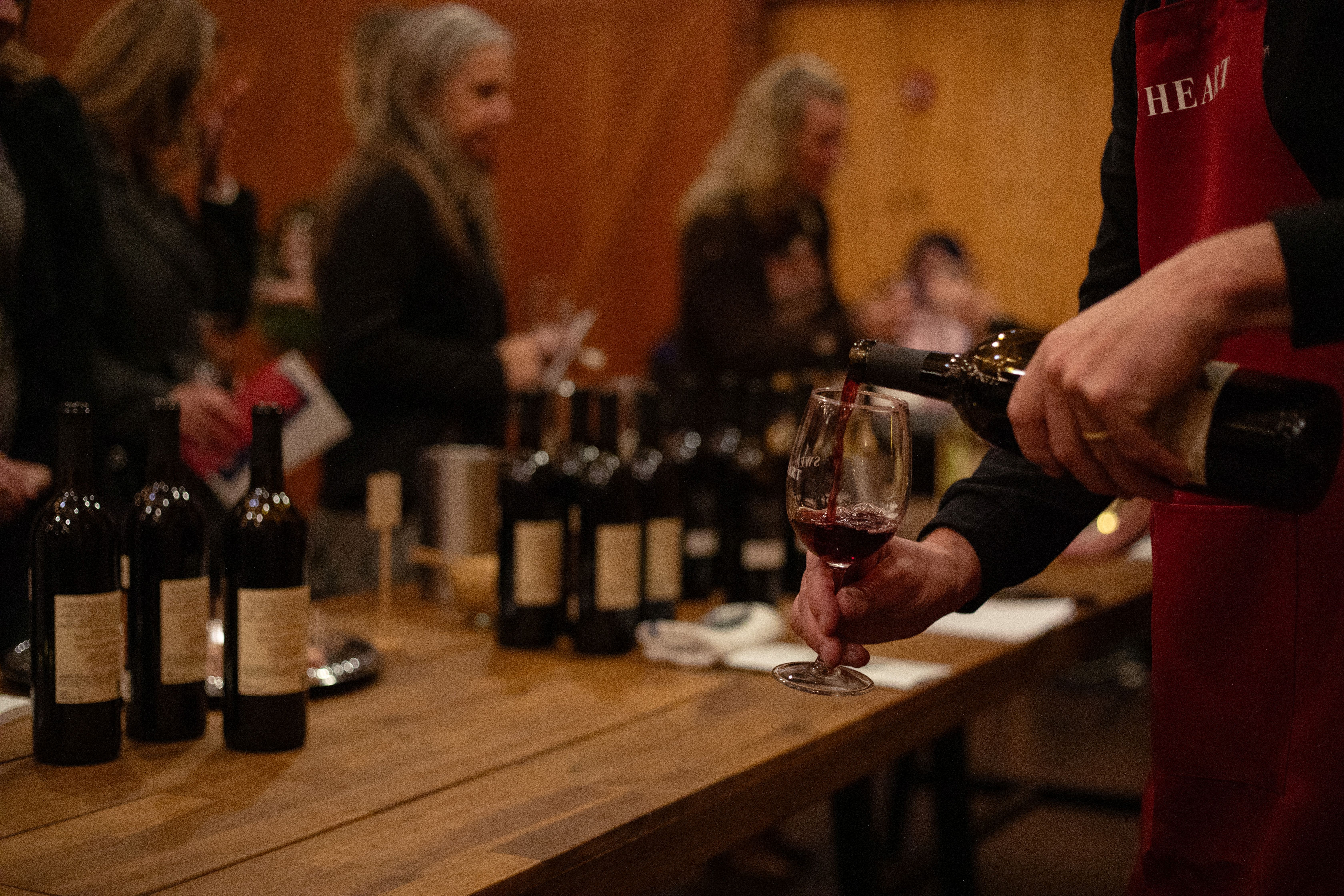 Person pouring red wine into a glass at a table with wine bottles, with people in the background.