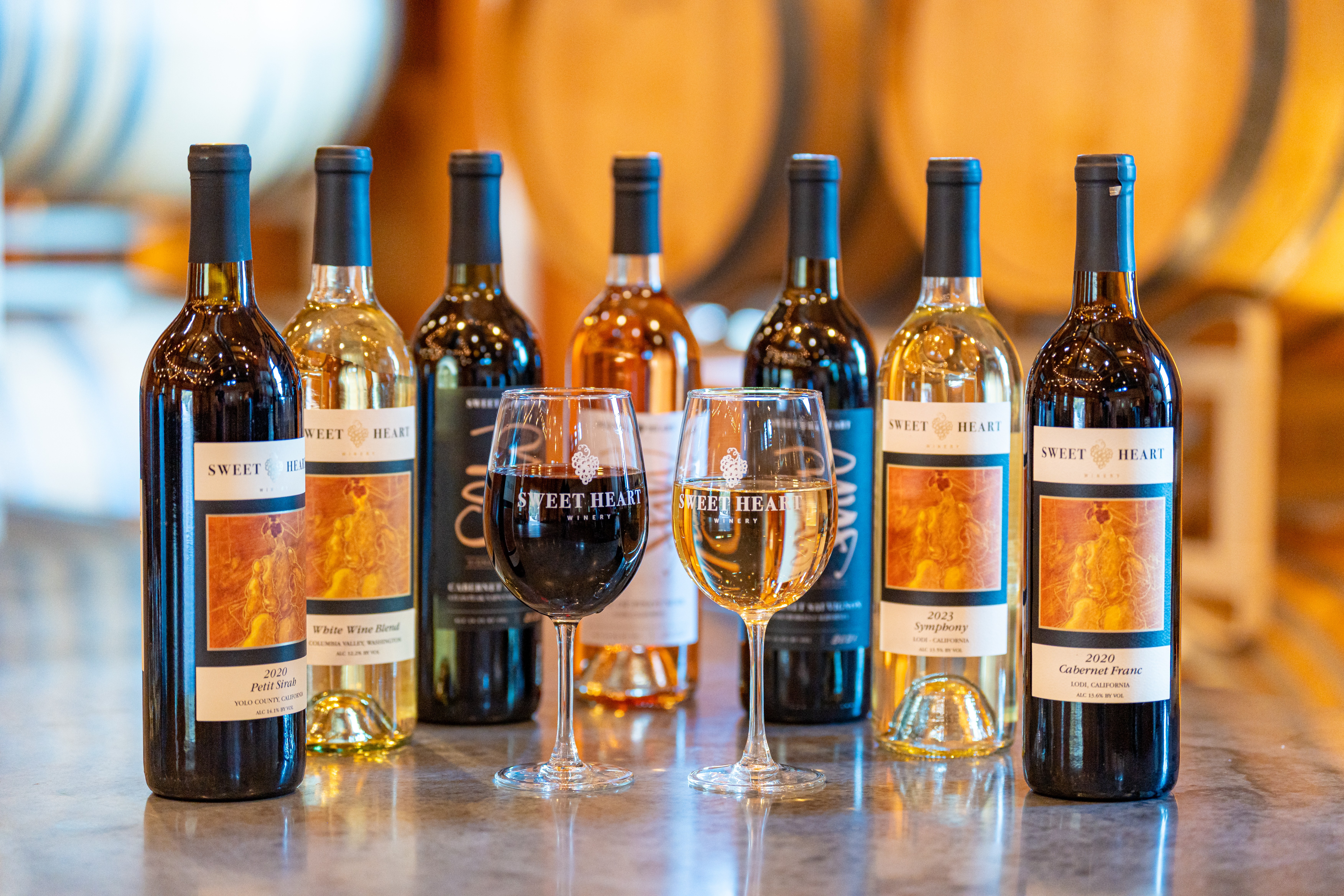 Seven wine bottles and two glasses of wine on a table with barrels in the background.