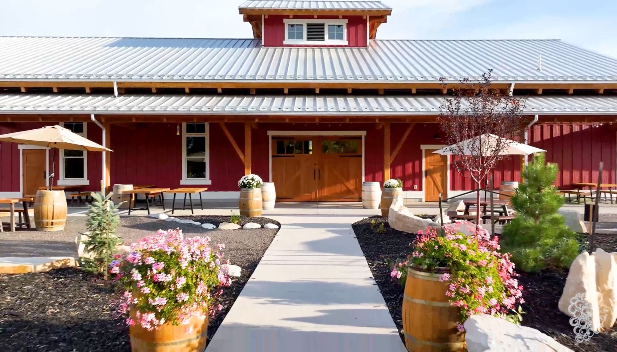 Red barn-style building with a metal roof, surrounded by potted plants and outdoor seating, under a clear sky.