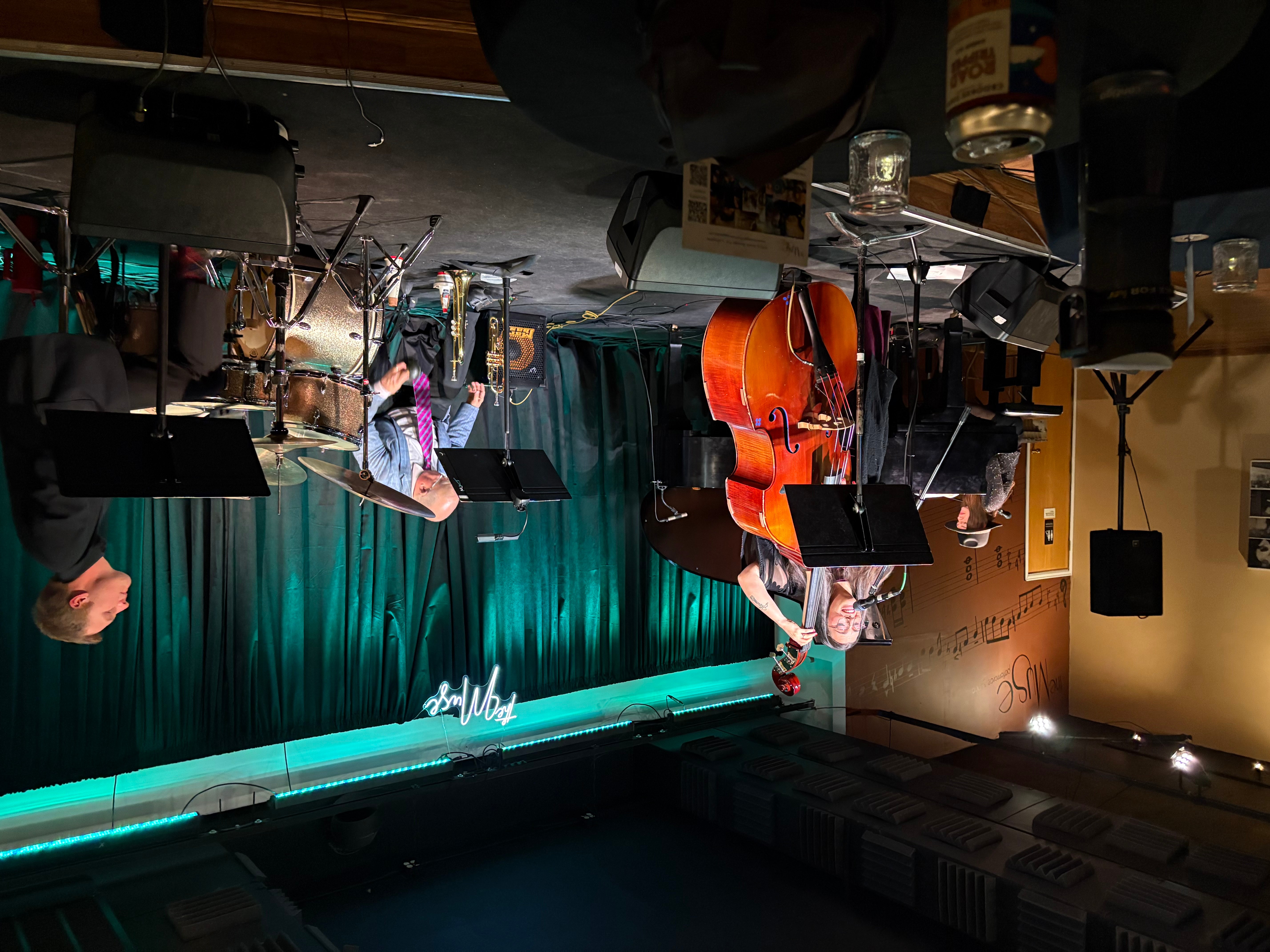 Musicians perform on stage with a teal curtain backdrop and a neon sign, playing instruments including a guitar and drums.
