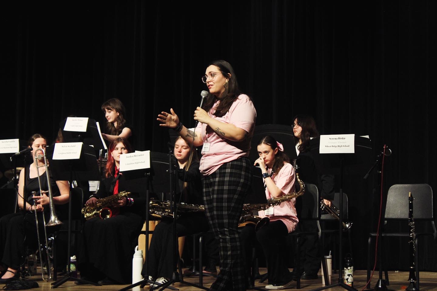Woman speaking into microphone on stage with seated musicians behind her, black curtain backdrop, some chairs empty