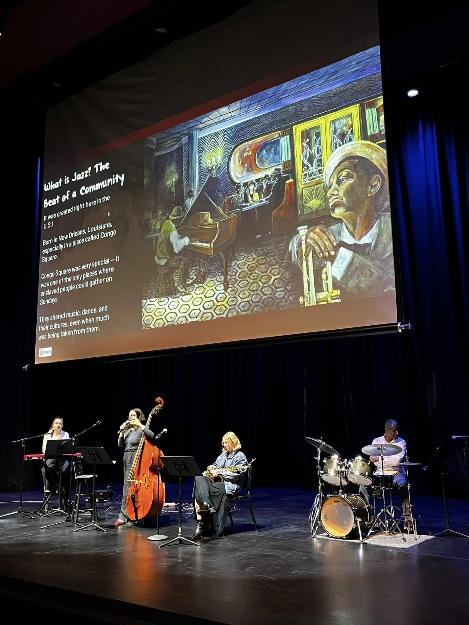 Four musicians perform on stage with a large screen displaying a painting of a room with people and artwork in the background.
