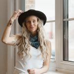 Woman with long wavy hair wearing a hat and scarf, standing by a window, looking at the camera.