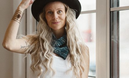 Woman with long wavy hair wearing a hat and scarf, standing by a window, looking at the camera.