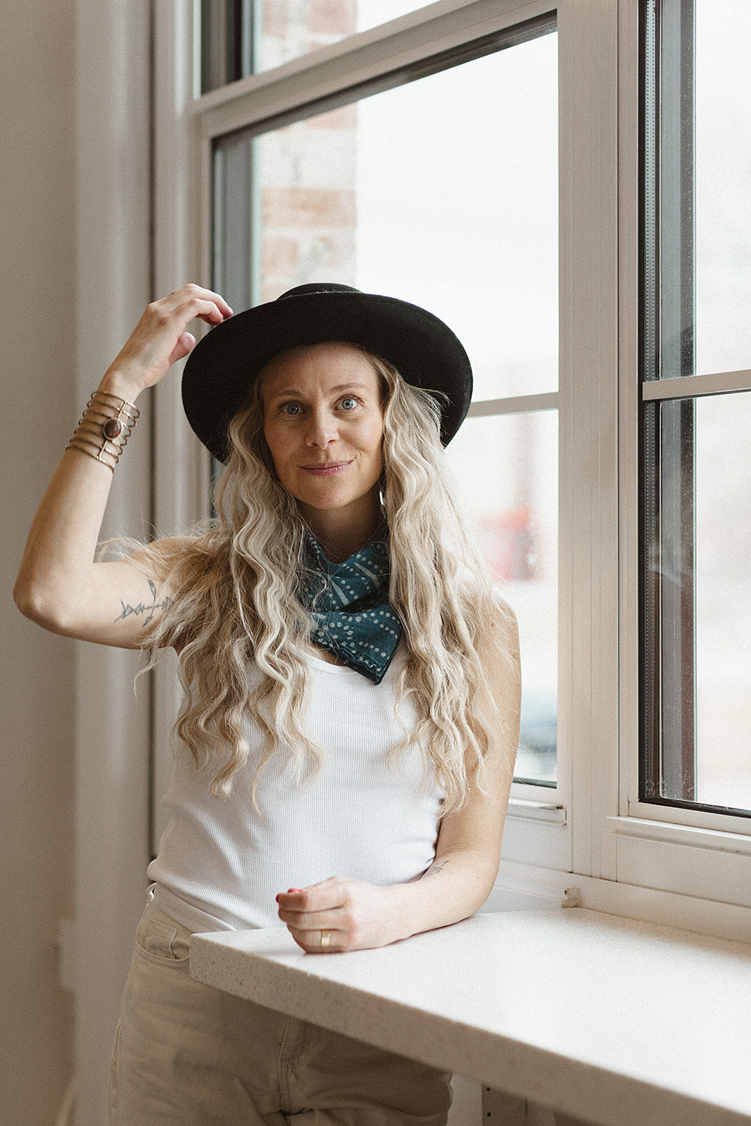 Woman with long wavy hair wearing a hat and scarf, standing by a window, looking at the camera.