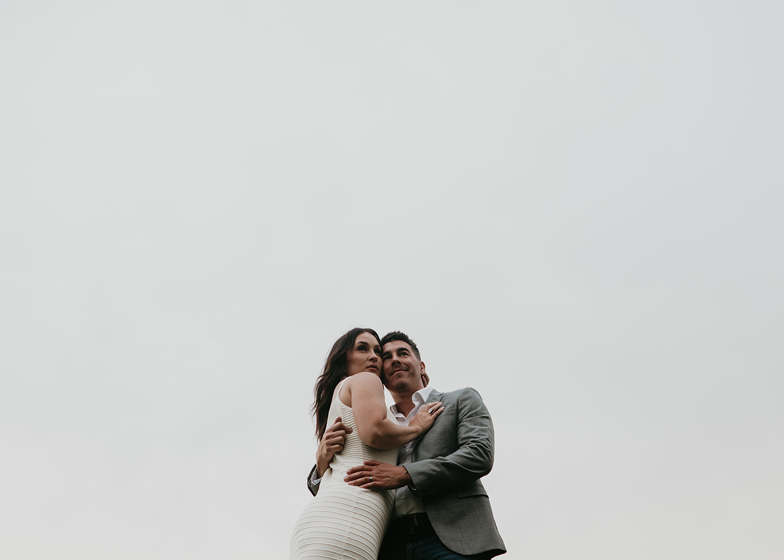 A couple stands close together, embracing, against a plain, light gray sky background.