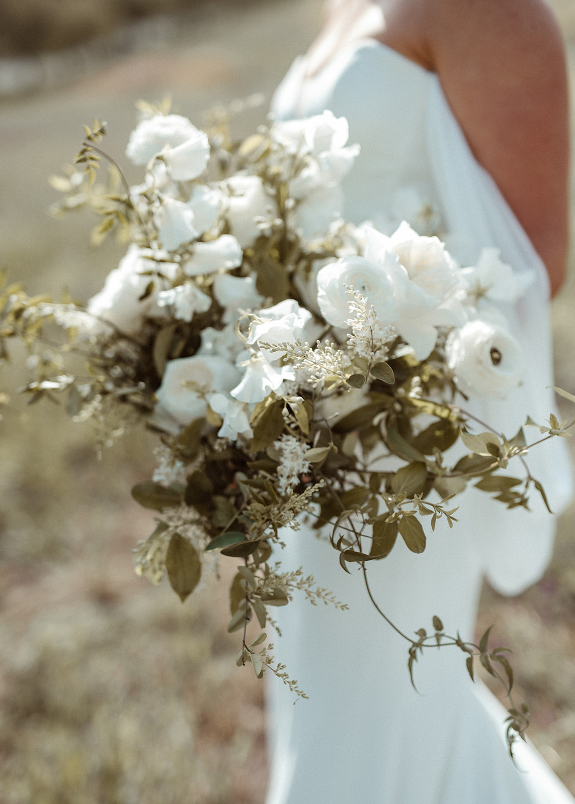 Person holding a large bouquet of white flowers and greenery, wearing a white dress, outdoors.