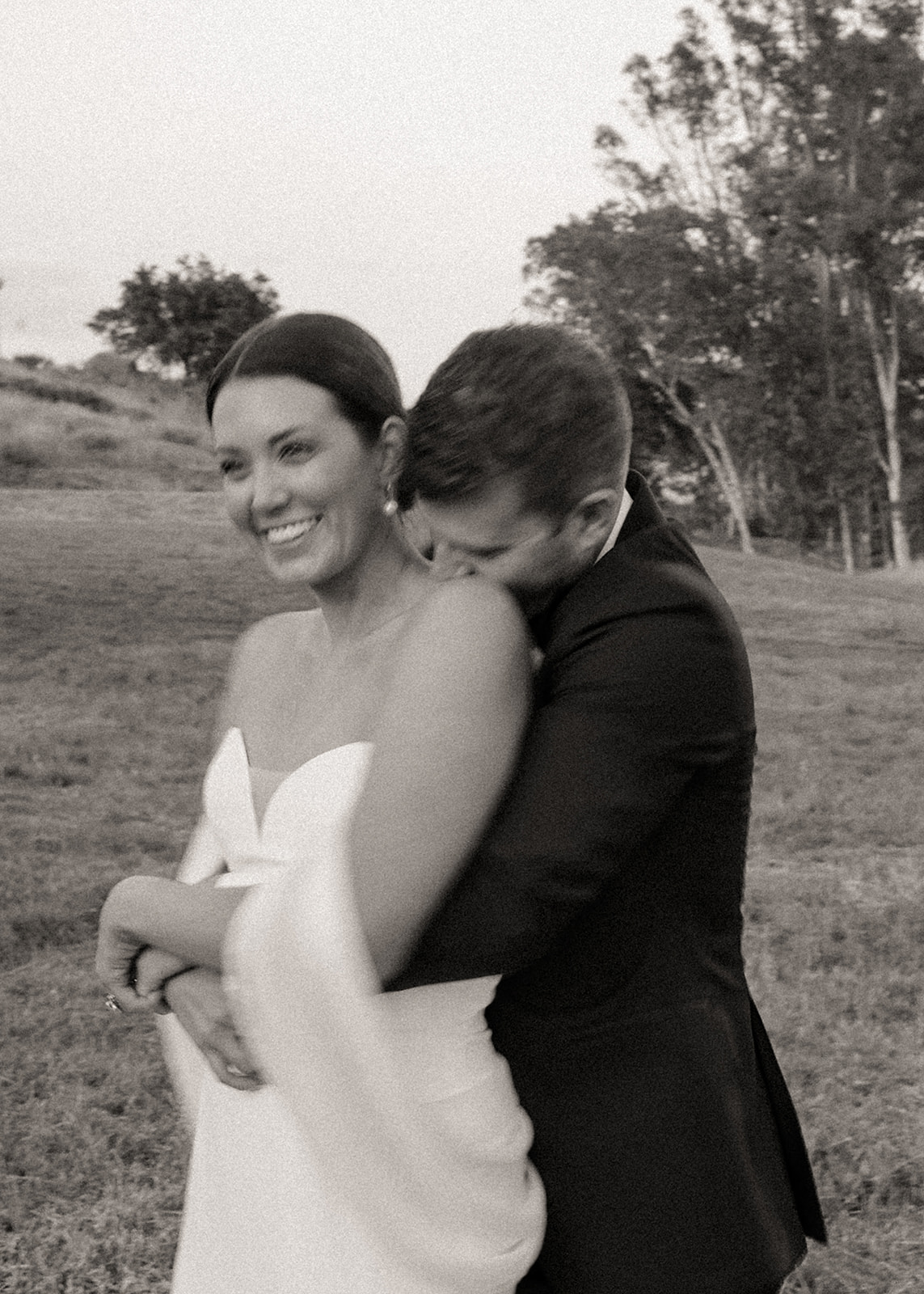 A smiling woman in a white dress is embraced by a person in a dark suit outdoors, trees in background.