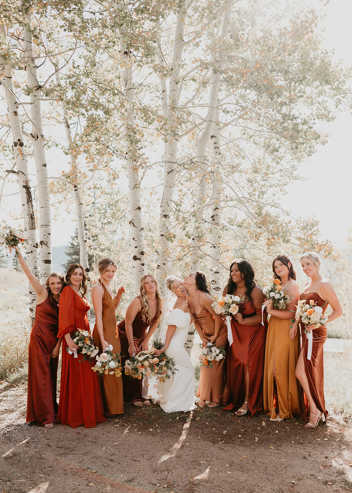 Group of women in colorful dresses and a bride in white, holding bouquets, standing outdoors with trees in background.