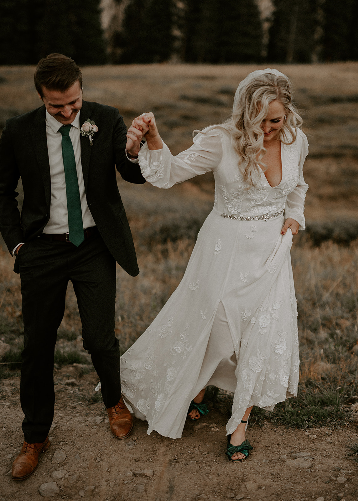 A couple holding hands, walking outdoors on a dirt path, with trees in the background. The woman wears a white dress, the man a suit.