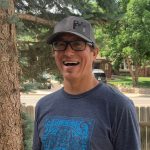 Smiling man wearing glasses and a cap outdoors, trees and a fence in background.