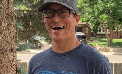 Smiling man wearing glasses and a cap outdoors, trees and a fence in background.