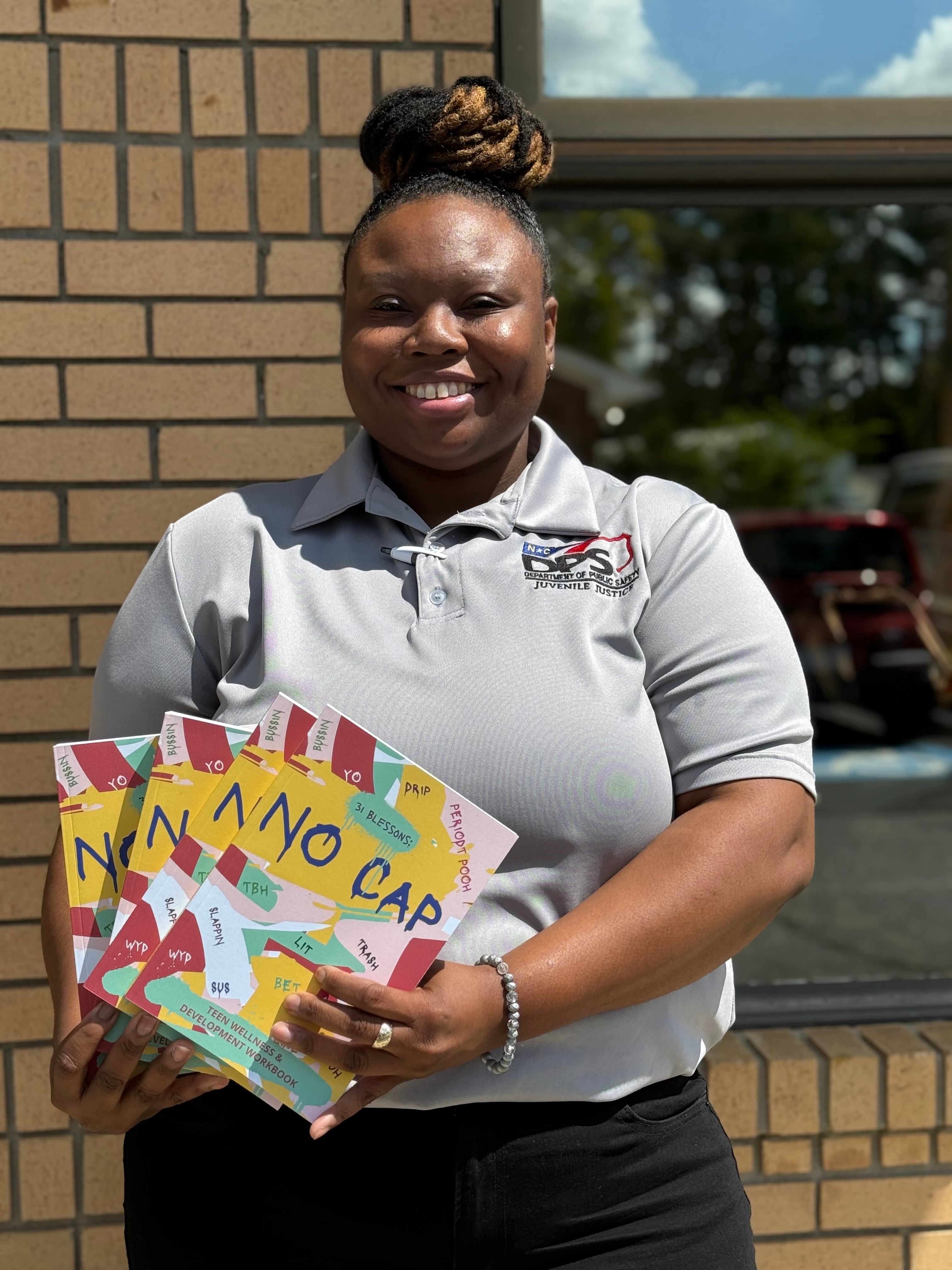 Smiling woman holding colorful books outside a building with a brick wall and window, sunny day.