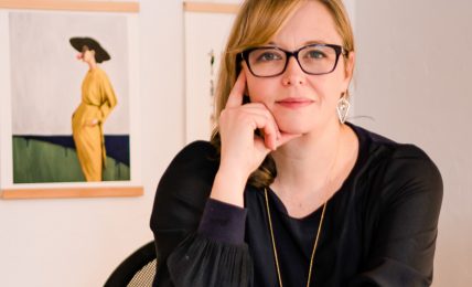 Woman with glasses sitting at a desk, resting her chin on her hand, with framed photos on the wall behind her.