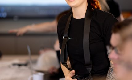 Smiling woman with red hair holding a camera in a meeting room, with a large screen in the background.
