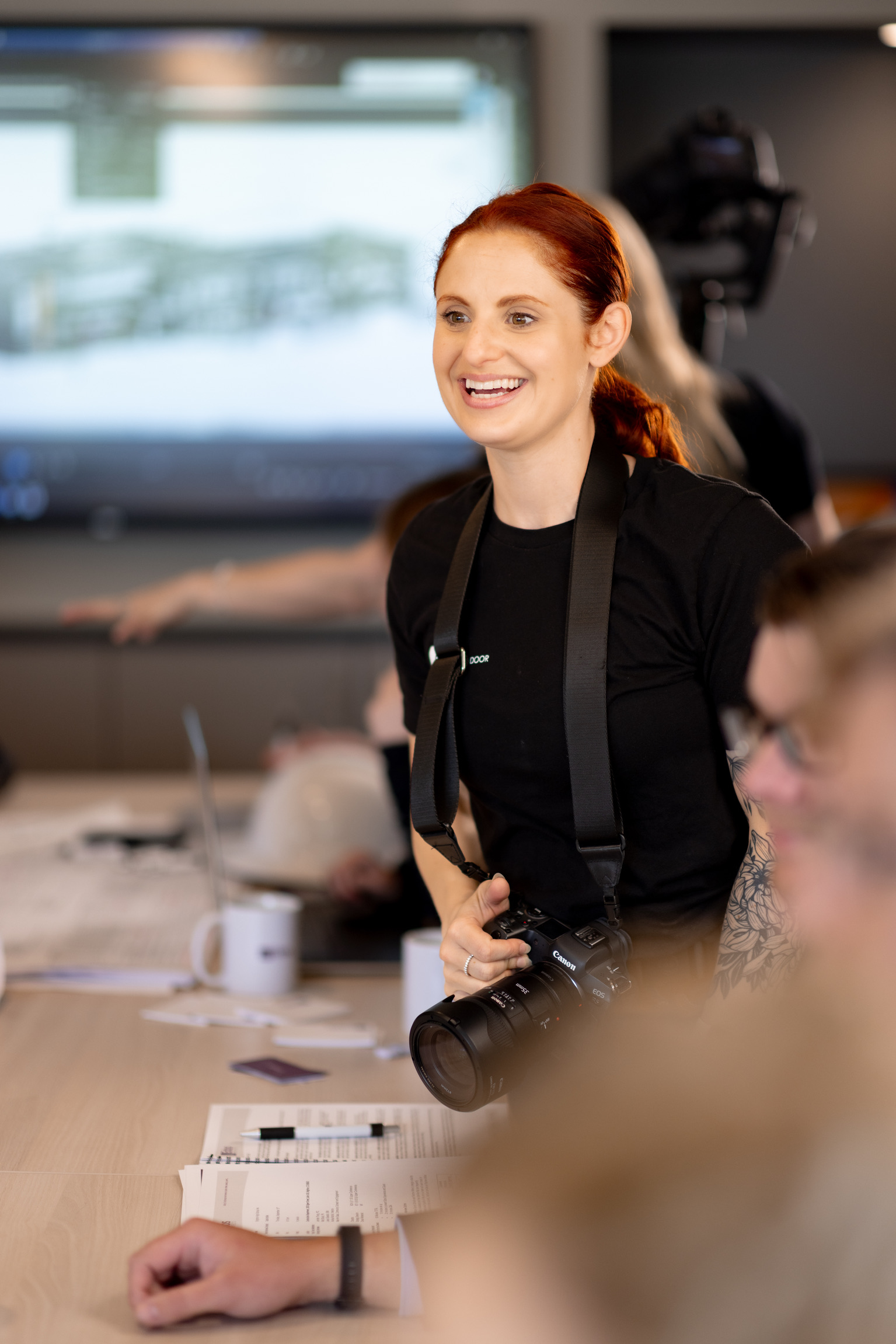 Smiling woman with red hair holding a camera in a meeting room, with a large screen in the background.