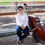 Person sitting on a park bench with a double bass, smiling, outdoors with trees and a fence in background.